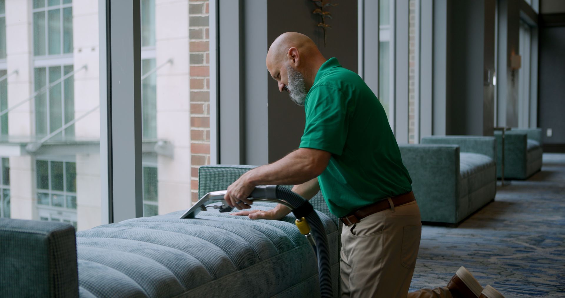 Man in green shirt cleans a blue bench with a vacuum. Large windows in the background.