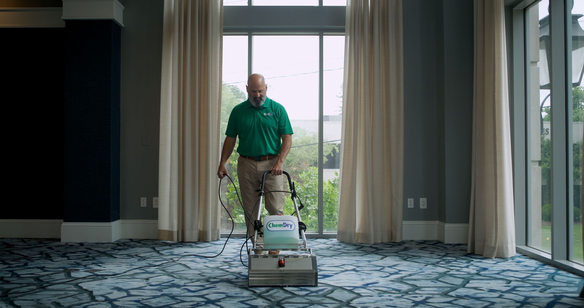 Man in green shirt using carpet cleaner on patterned blue carpet in large room with large windows.