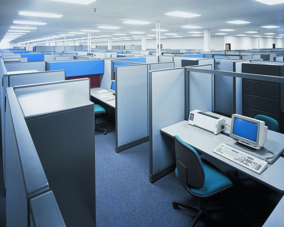 Empty office cubicles with blue and gray partitions, blue carpet, and overhead lights.