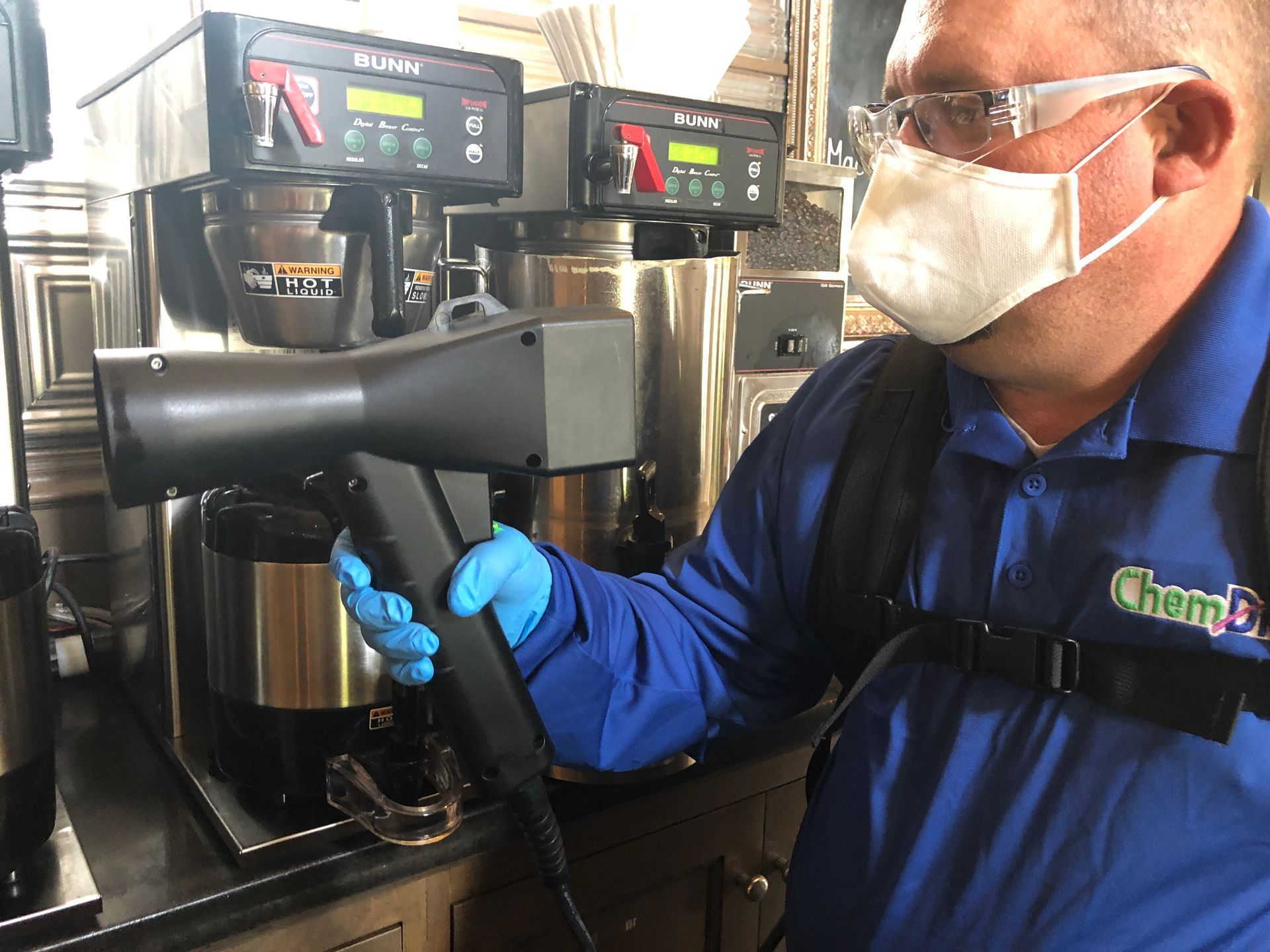 Person in blue uniform, mask, and gloves disinfecting coffee machines with a spray tool.