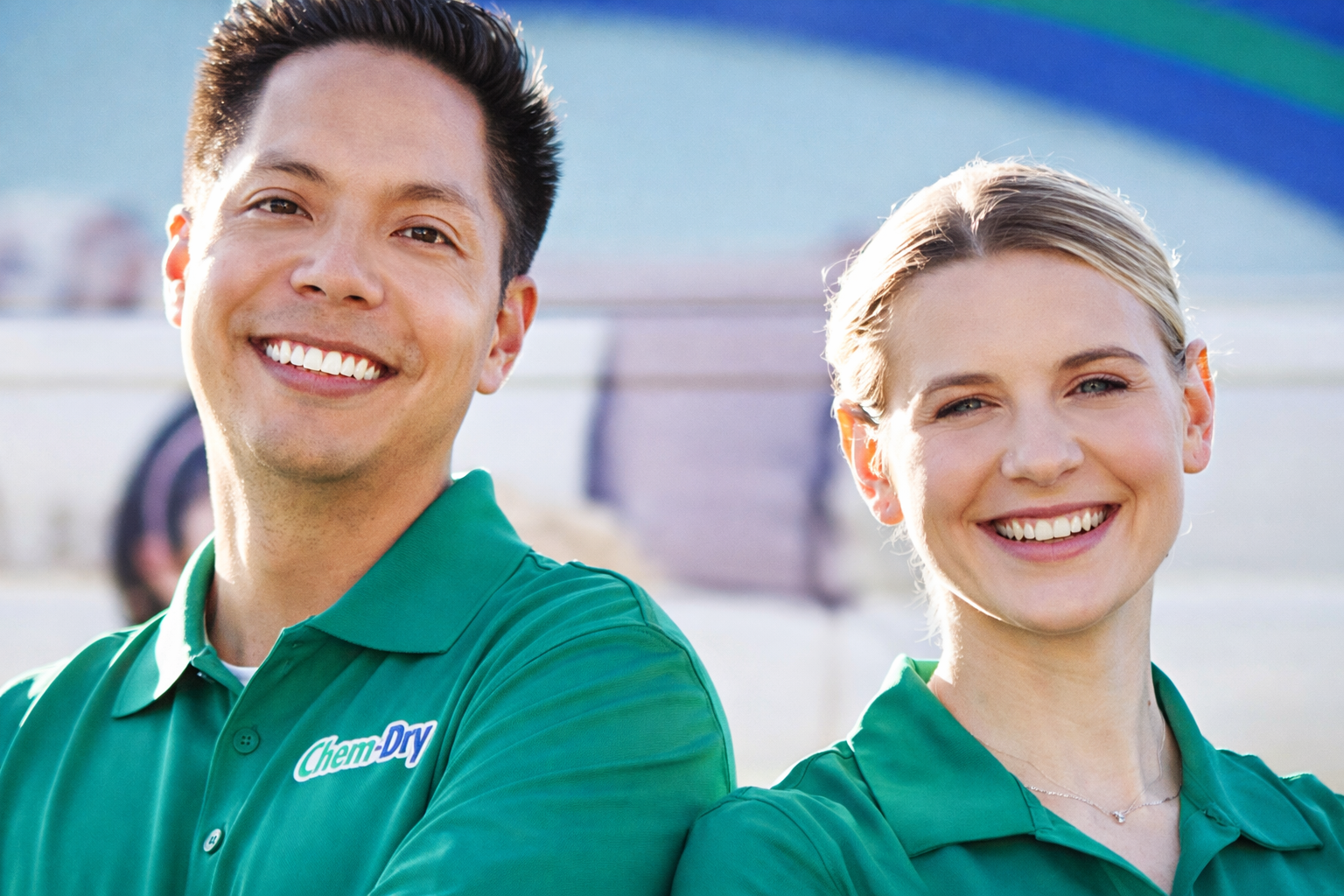 Two smiling people in green shirts stand outside by a vehicle.