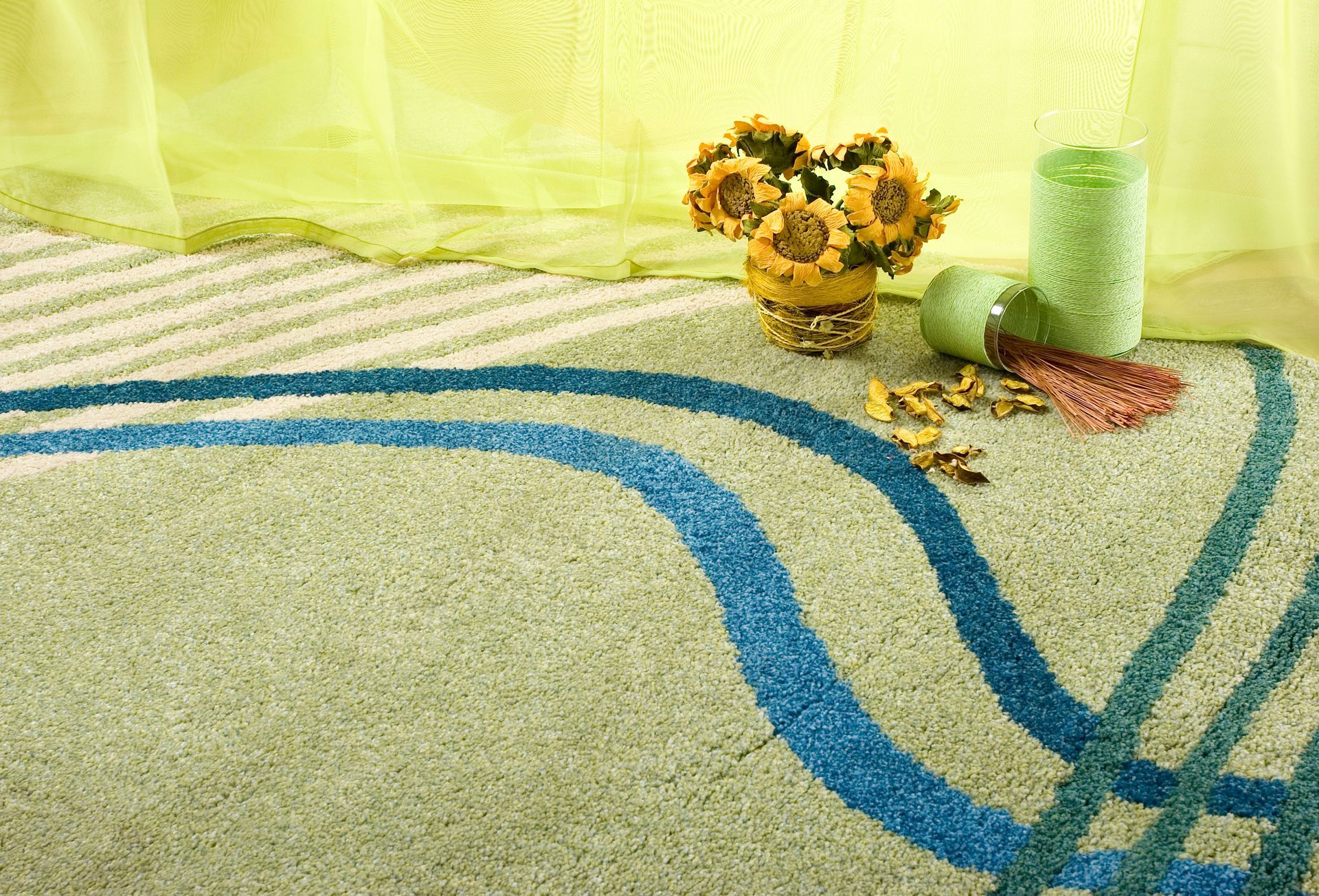 A rug with blue lines, sunflowers in a vase, and a green candle with a light green curtain in the background.