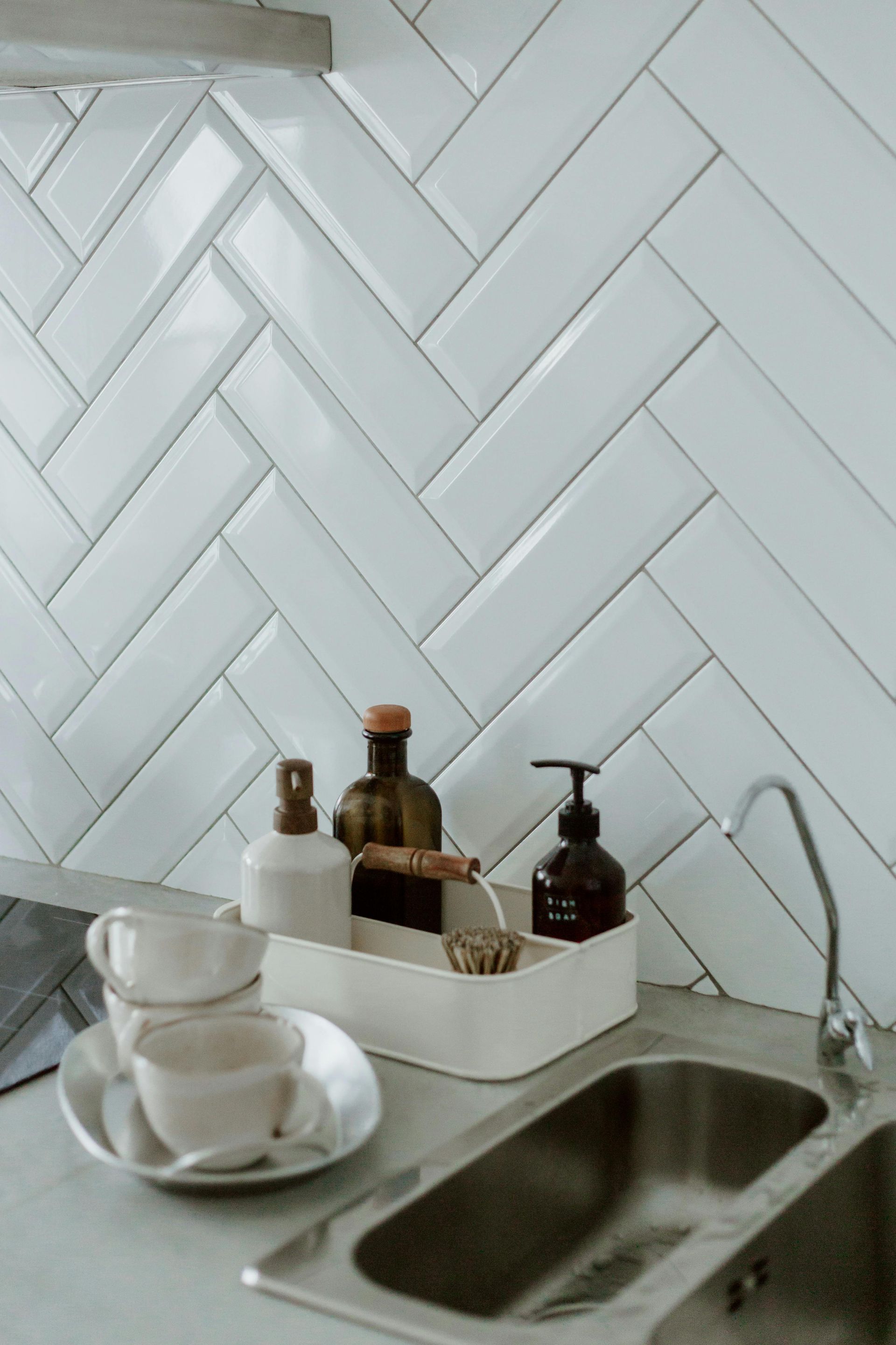 Kitchen sink with white herringbone backsplash, bottles, cups, and a brush.