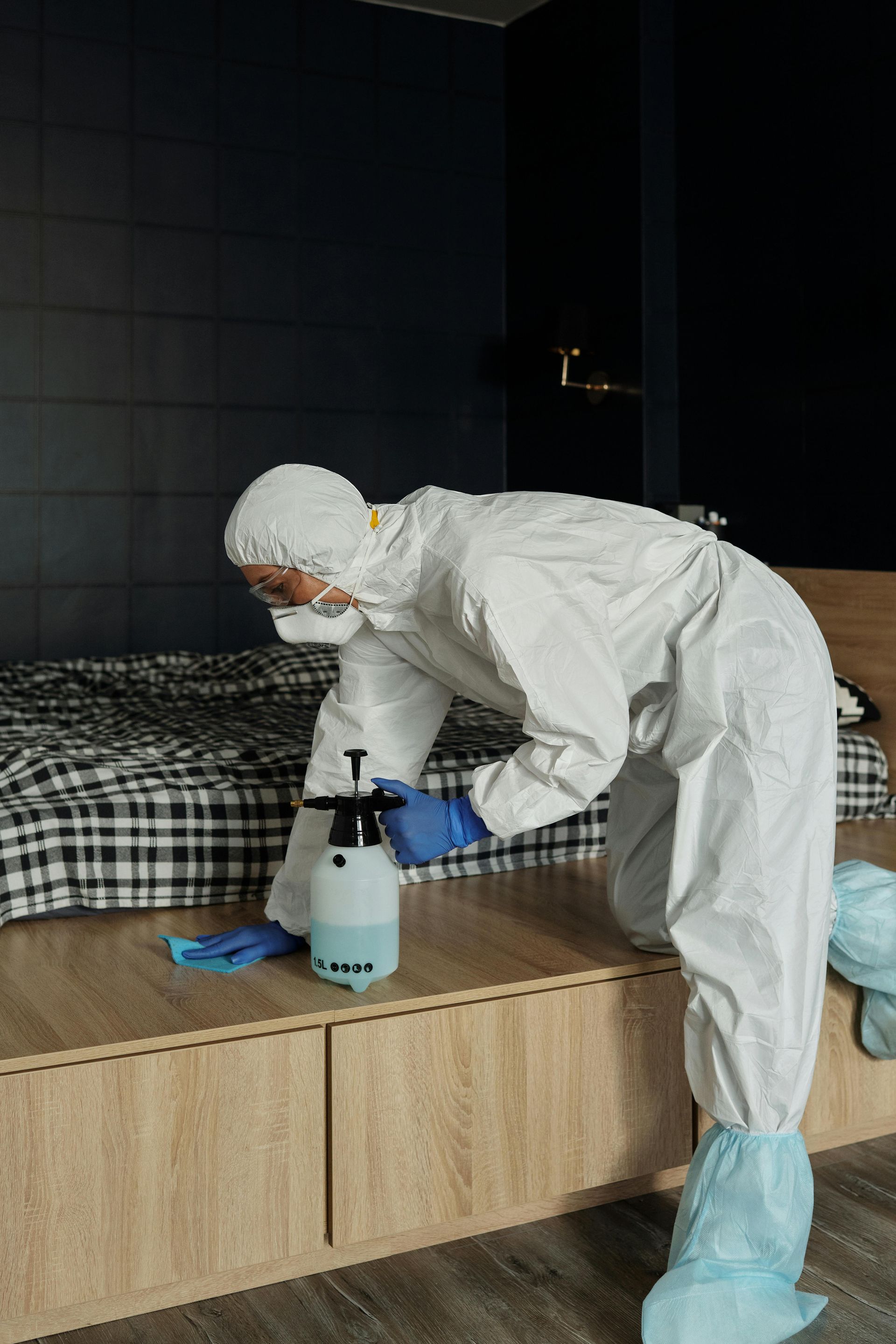Person in protective suit disinfecting a bedroom with a spray bottle and cloth.