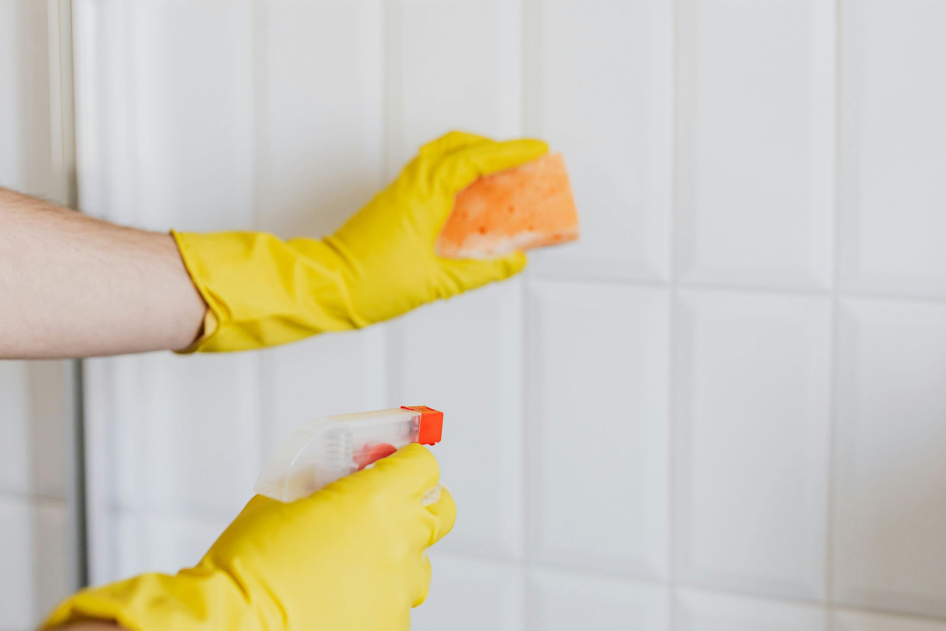 Hands in yellow gloves cleaning white tiled wall with spray bottle and sponge.