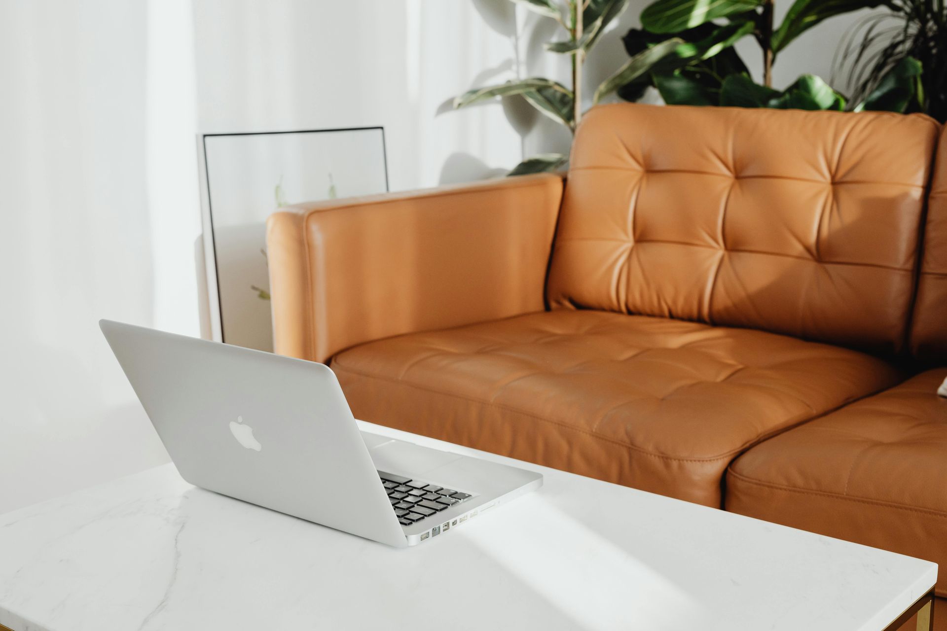 Laptop on a white table in front of a brown leather couch in a bright room.