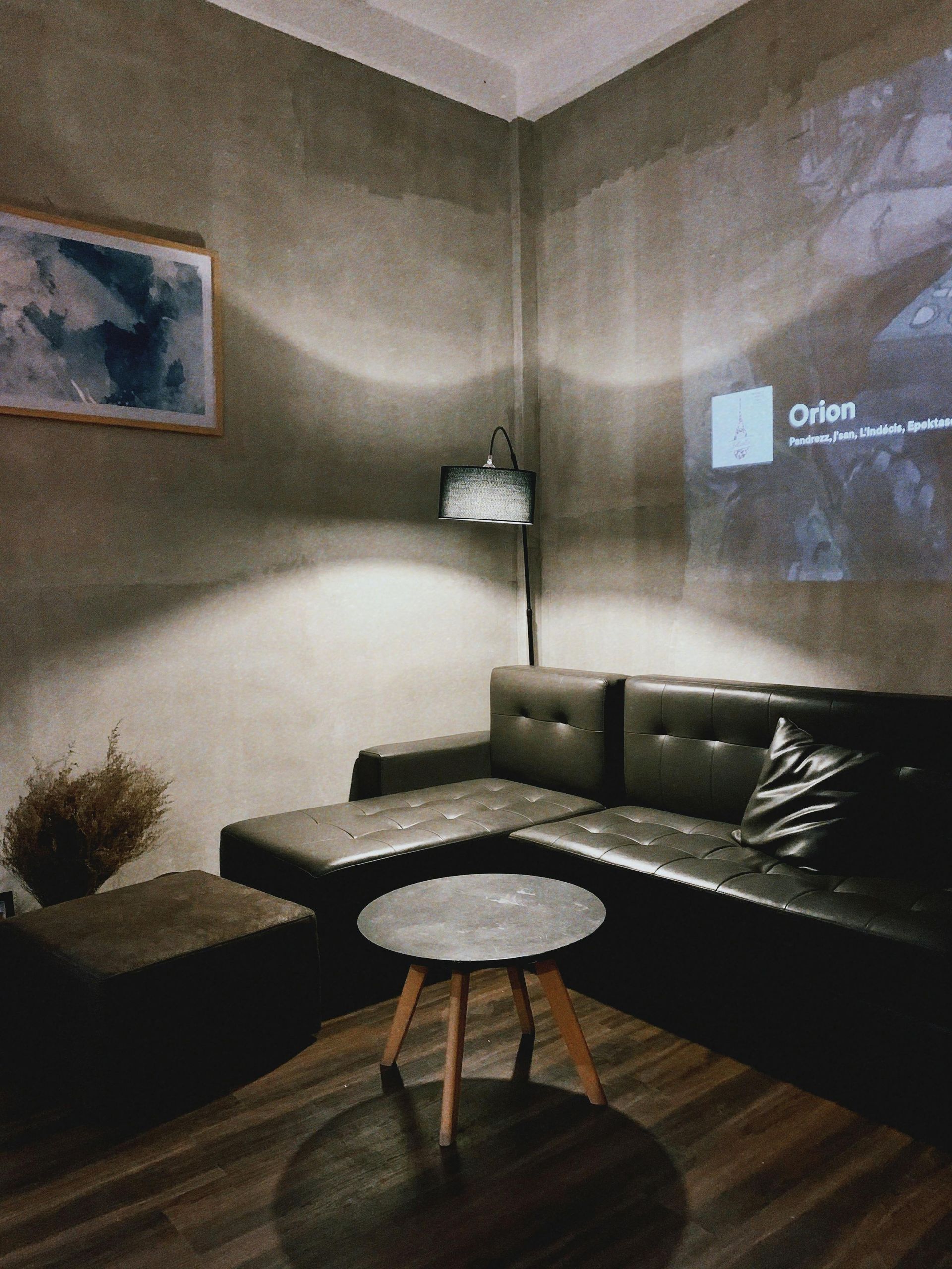 Cozy room with a dark leather sectional sofa, small round table, and a projector screen. Concrete walls and dim lighting.