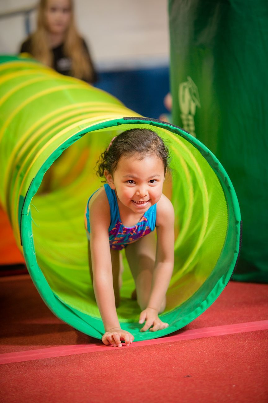 Girl crawls through a green and yellow tunnel on a red floor. She smiles and wears a leotard.