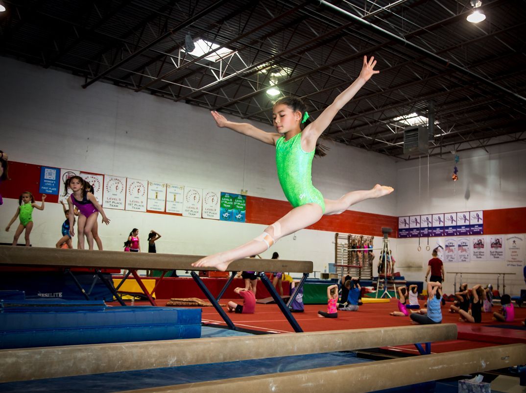 Young gymnast in green leotard leaps over balance beam in a gym; other children watch.