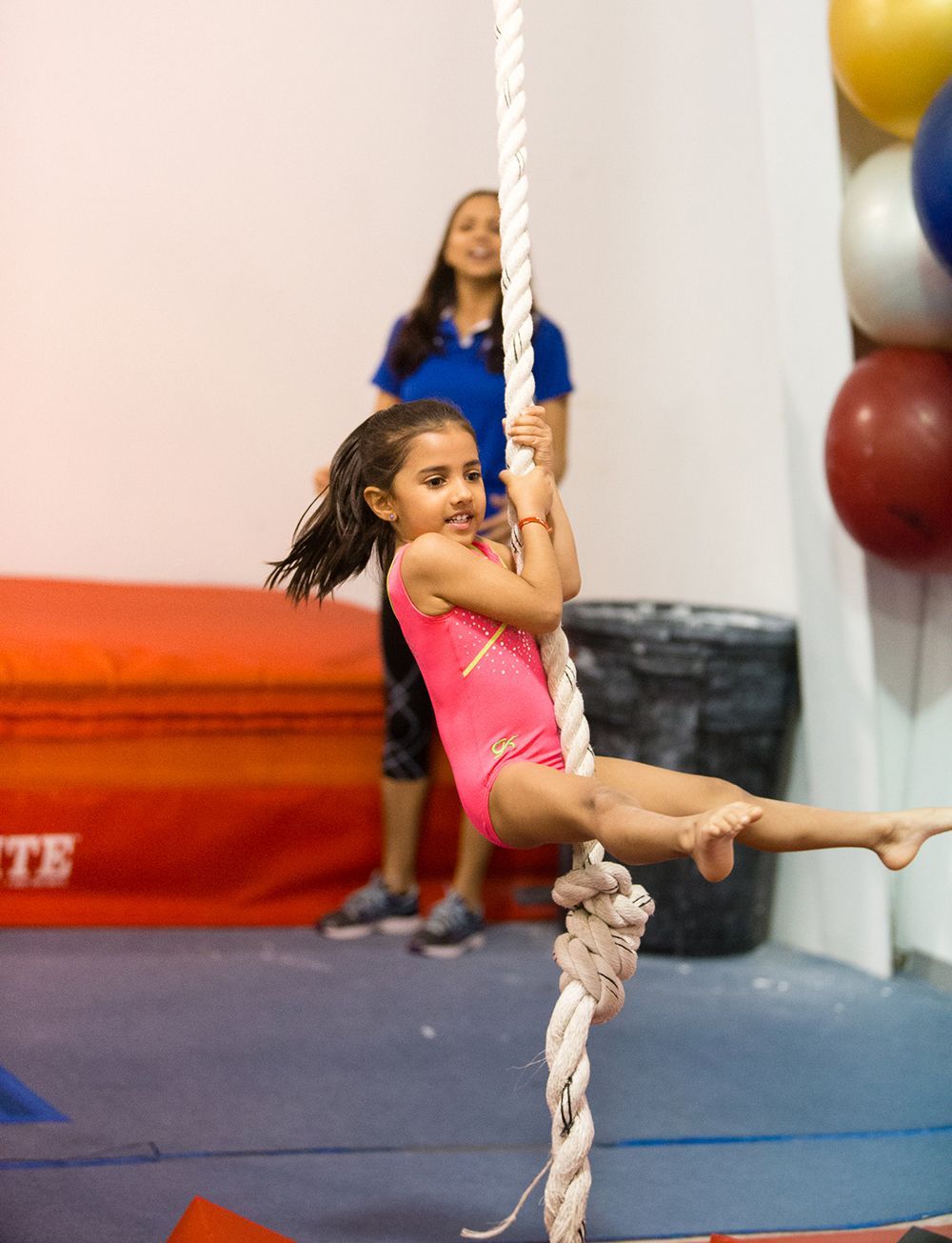 Young girl in pink leotard hangs from a rope at a gym, smiling. A woman watches in the background.