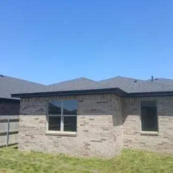 Brick house exterior with a dark roof and windows, viewed from a grassy backyard under a blue sky
