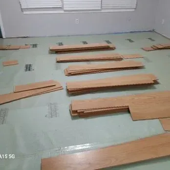 Wood flooring planks laid out across a room on top of green underlayment, ready for installation.