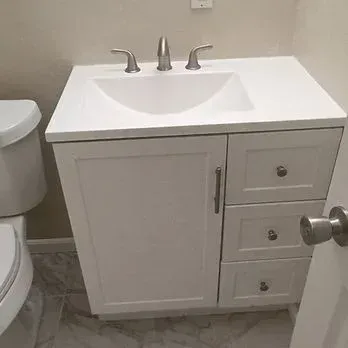 White bathroom vanity with sink, chrome faucet, and three drawers beside a toilet.