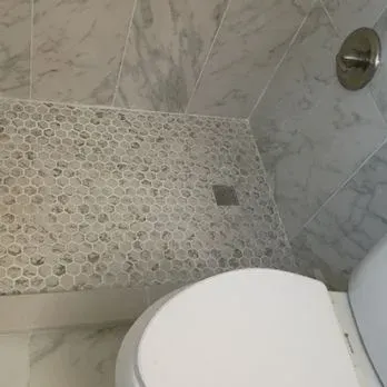 Small bathroom shower with pebble tile floor, white marble walls, and a toilet in the foreground