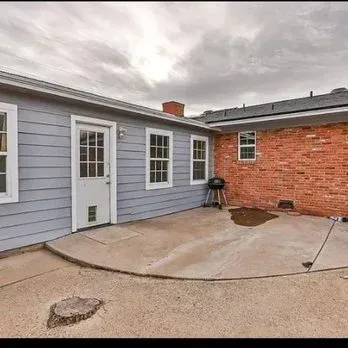 Small courtyard with gray siding, red brick wall, concrete patio, and a grill under cloudy sky.