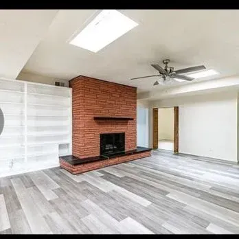 Empty living room with gray flooring, brick fireplace, ceiling fan, and skylight divider
