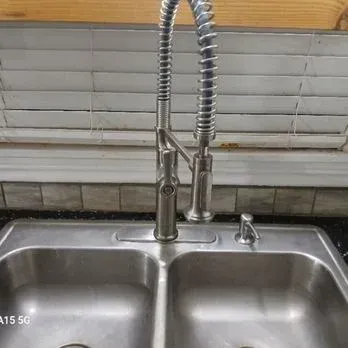Stainless steel kitchen sink with a spring-coil faucet and closed blinds behind it