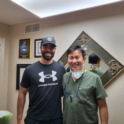 Two smiling people stand indoors, posing beside framed wall art and a diamond-shaped mirror.