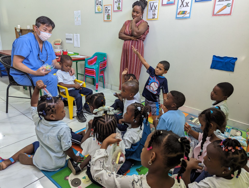 Teacher reading to seated children in a classroom, with one child raising a hand.