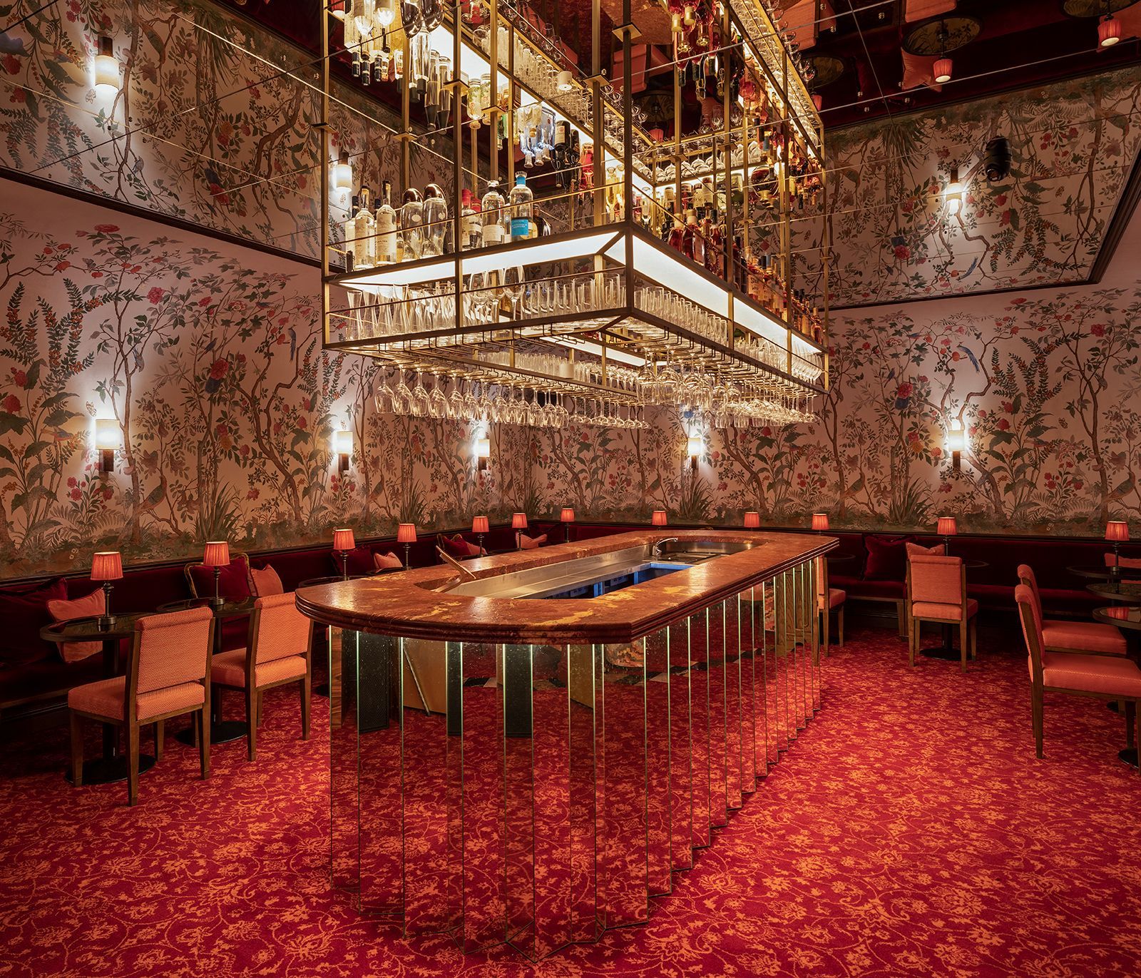 A luxurious bar with a mirrored counter, red carpet, floral wallpaper, and a suspended liquor rack.