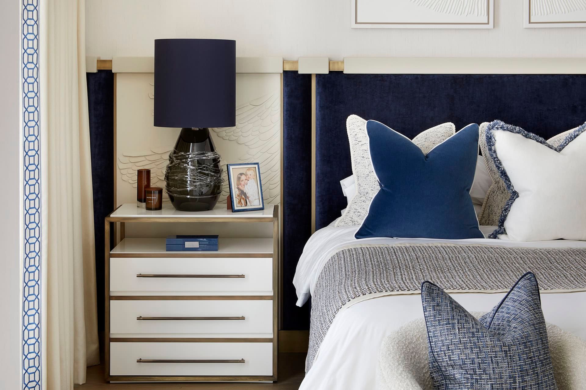 Bedroom with a blue velvet headboard, white bedding, and a nightstand with a blue lamp and picture frame.