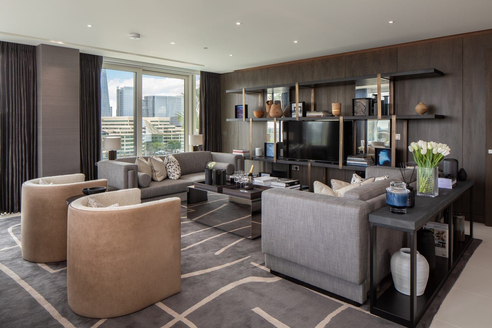 Modern living room with gray sofas, neutral armchairs, and a large wall-mounted TV against a dark wood wall.