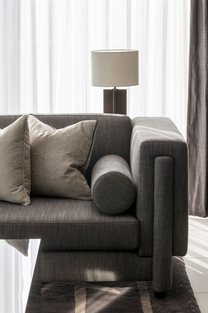 Gray upholstered sofa with decorative pillows, cylindrical bolster, and lamp beside a sheer curtain.