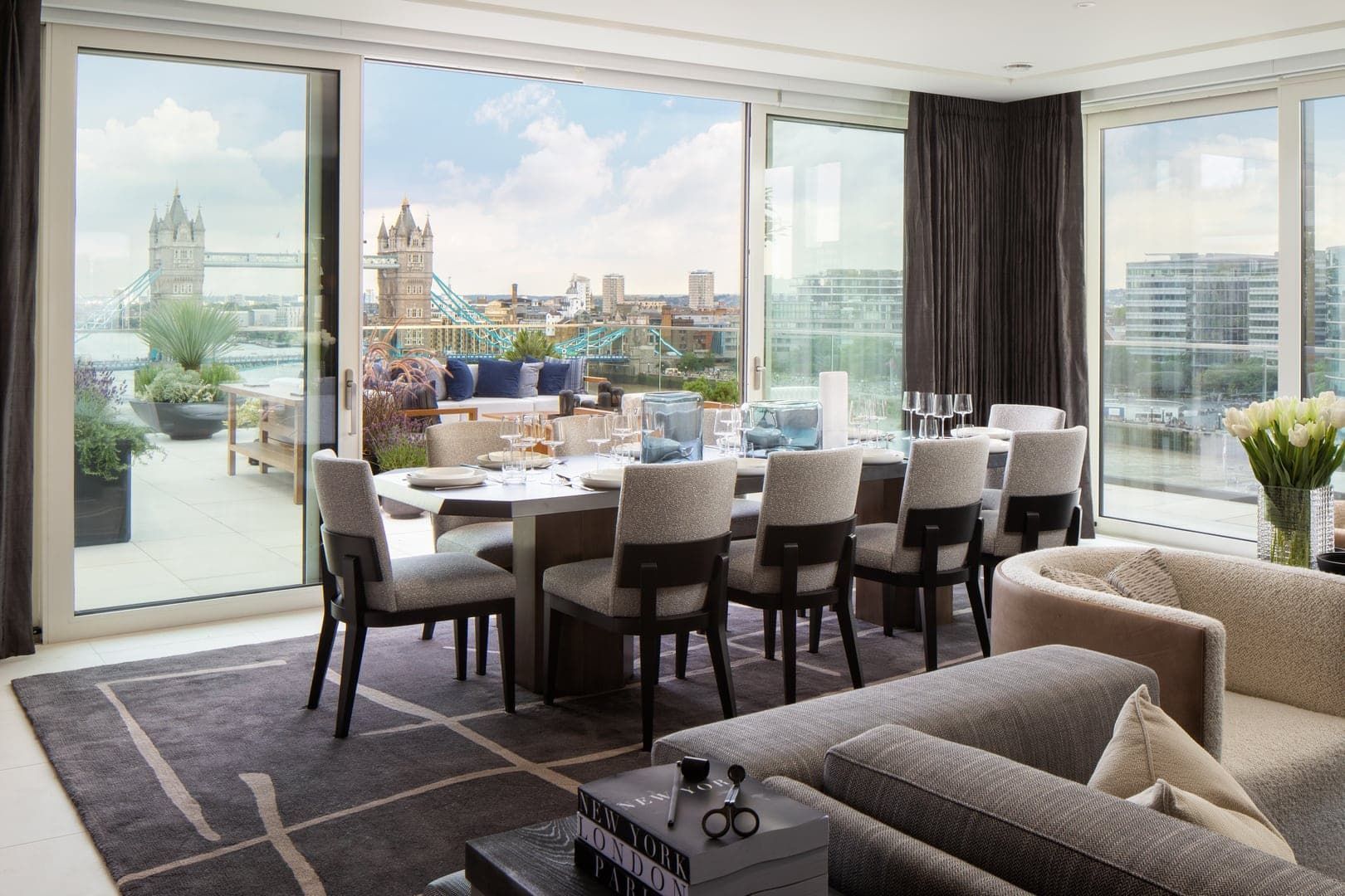 Dining room overlooking London's Tower Bridge. Table set for dinner, gray chairs, large windows, gray rug, neutral colors.