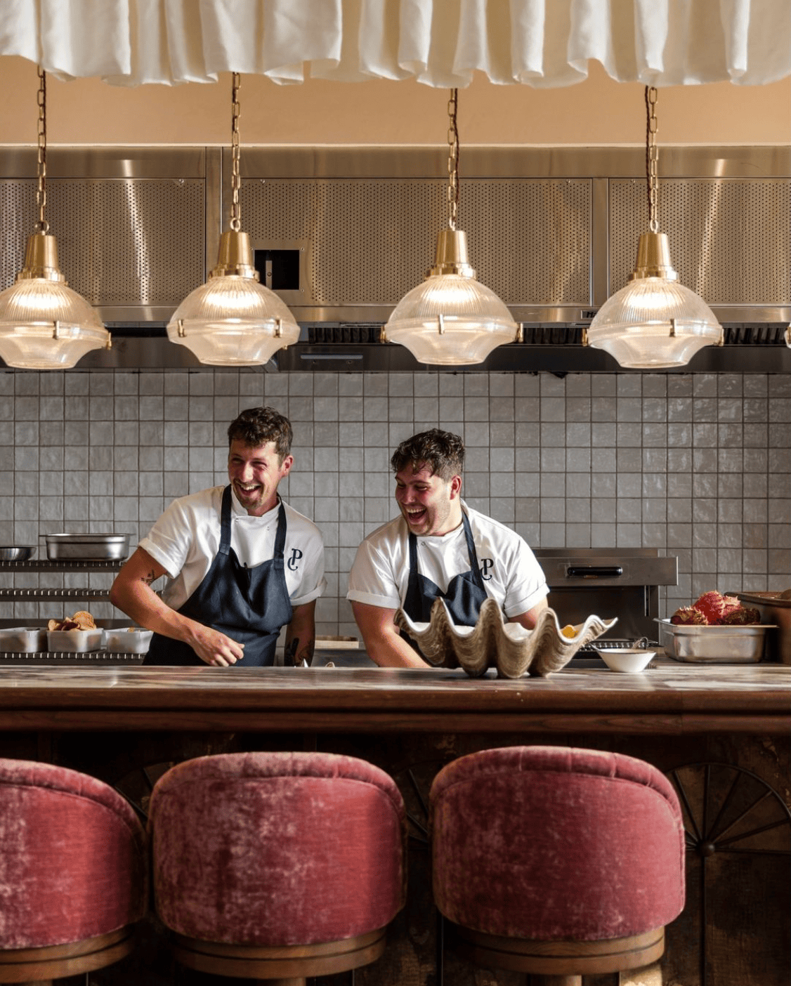 Two chefs laughing in a stylish kitchen, standing behind a counter with red stools.