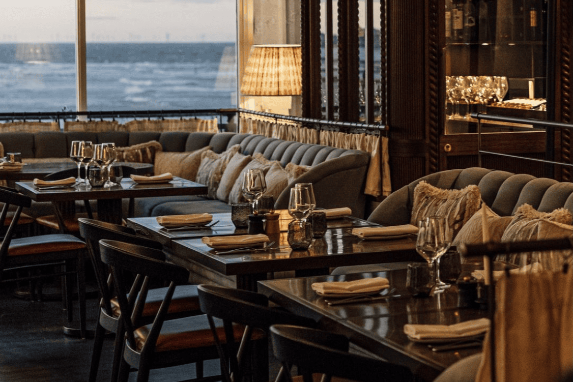 Restaurant dining area with tables, plush seating, ocean view, and warm lighting.