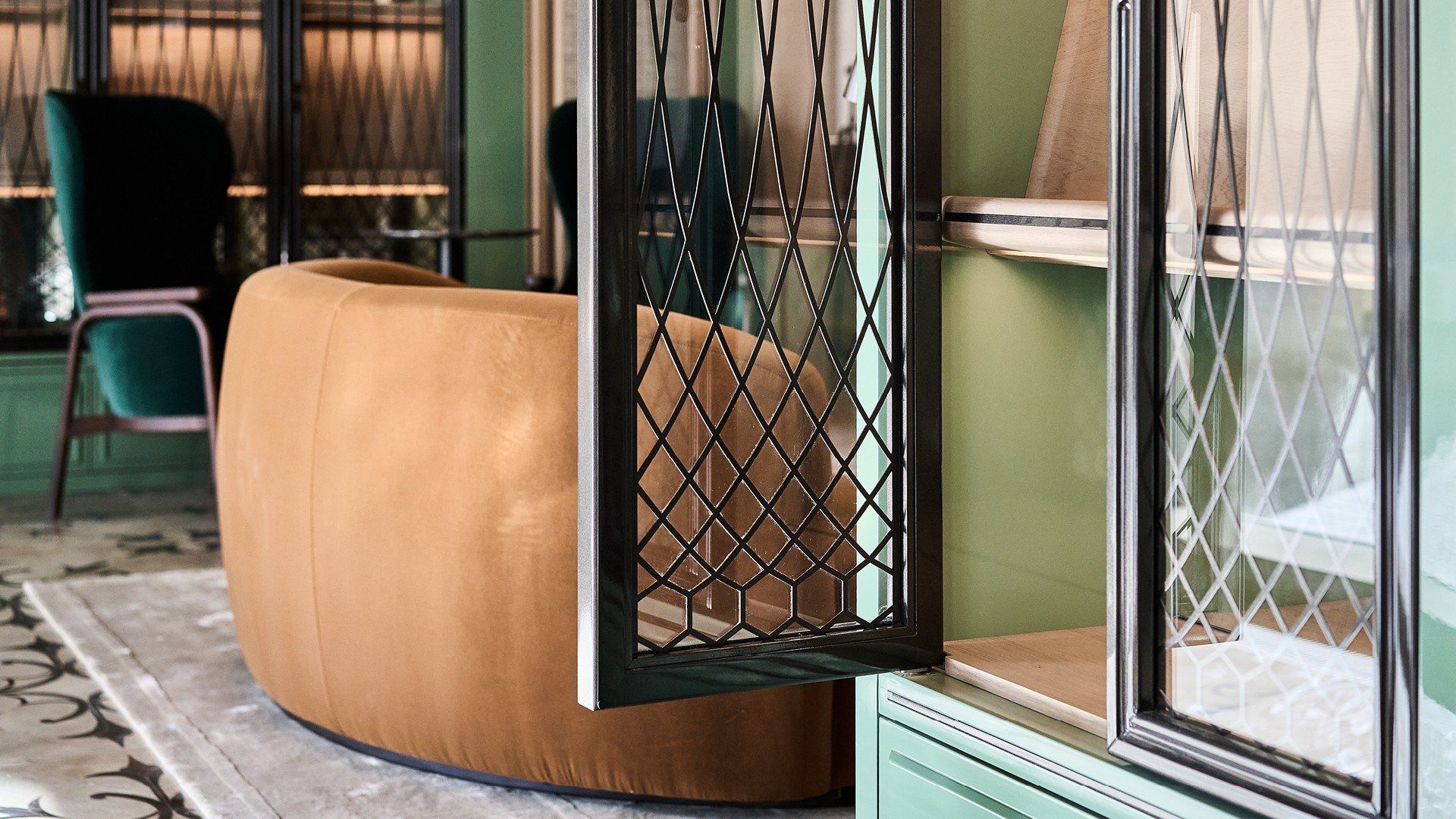 A curved tan couch, teal chair, and black latticed doors in a green room with patterned rug.