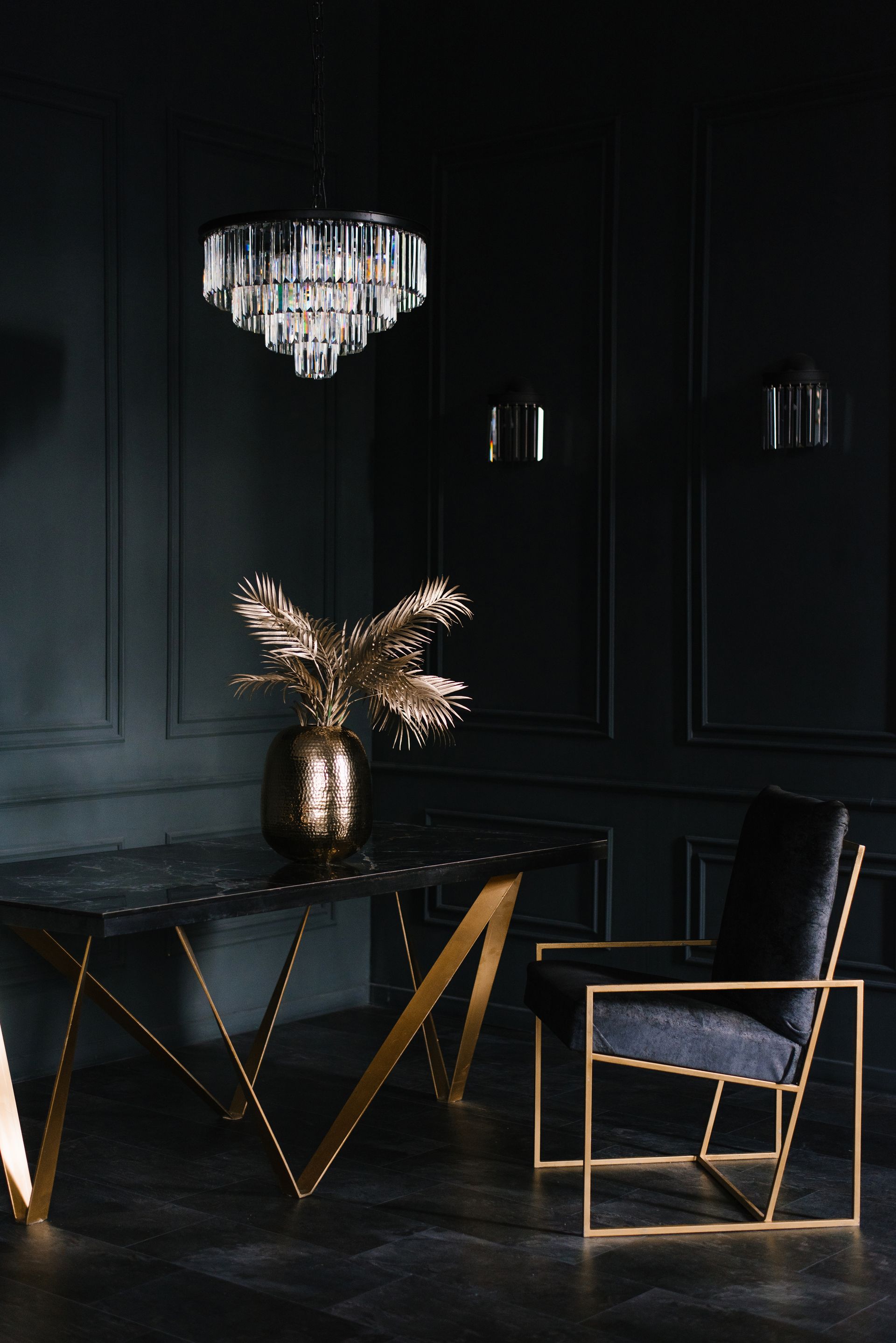 Dark room with gold accents: chandelier, table with gold legs, chair, vase with gold leaves.