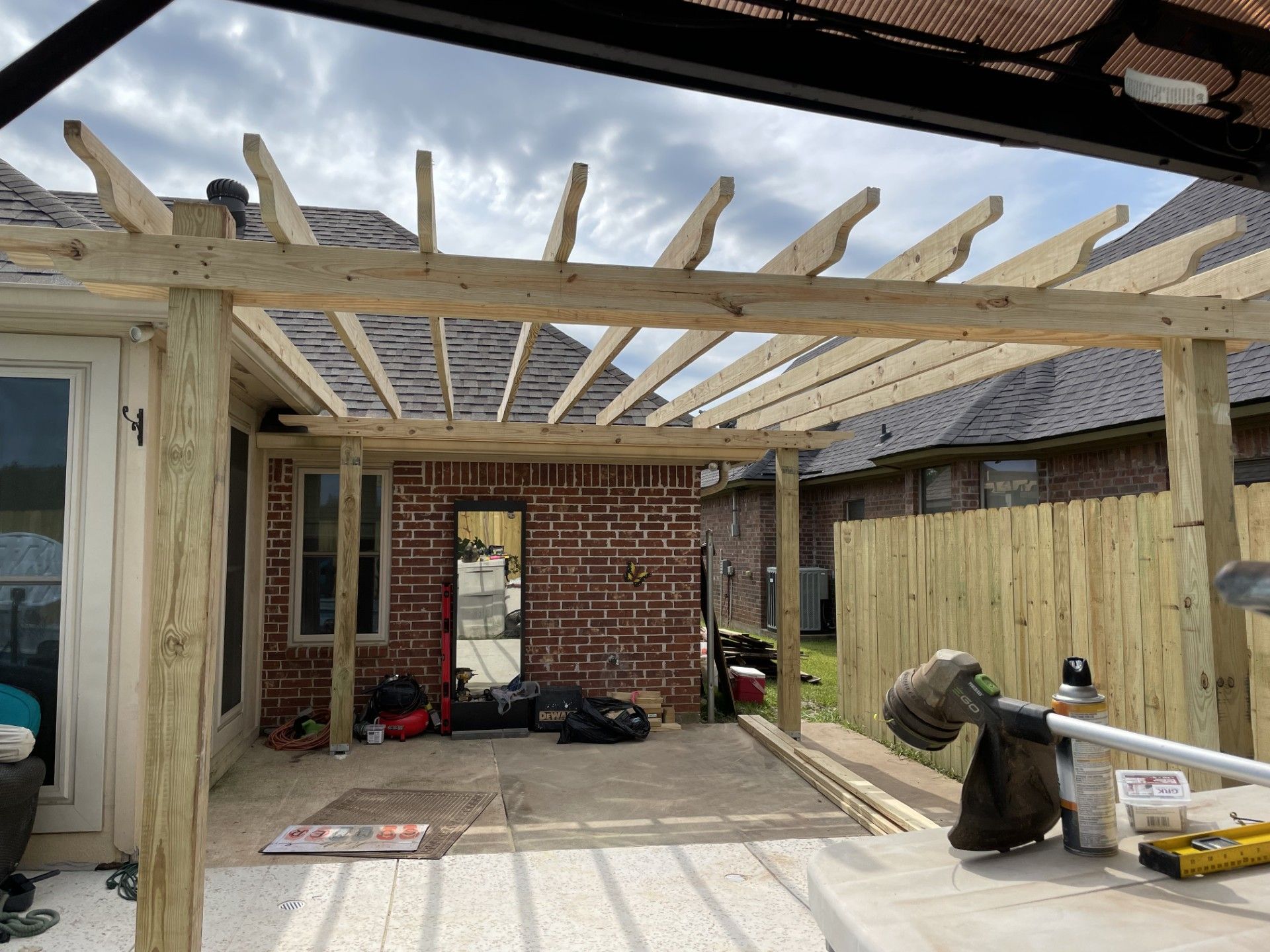 Wooden pergola under construction; patio with brick wall and fence in the background.