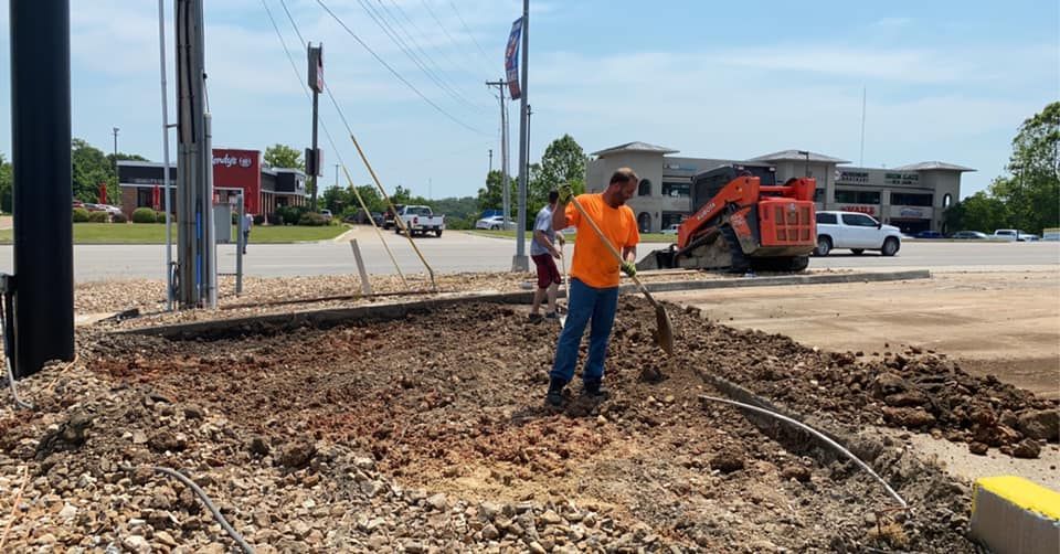 A yellow excavator is digging a hole in a parking lot in front of a building.