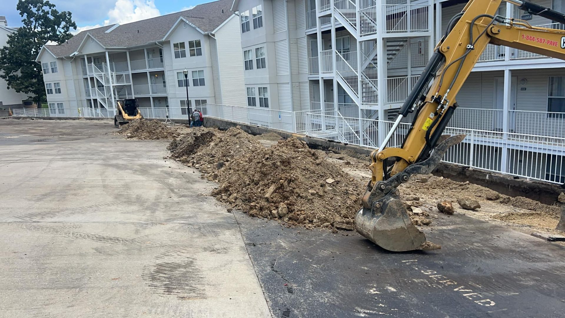 A yellow excavator is digging a hole in a parking lot in front of a building.