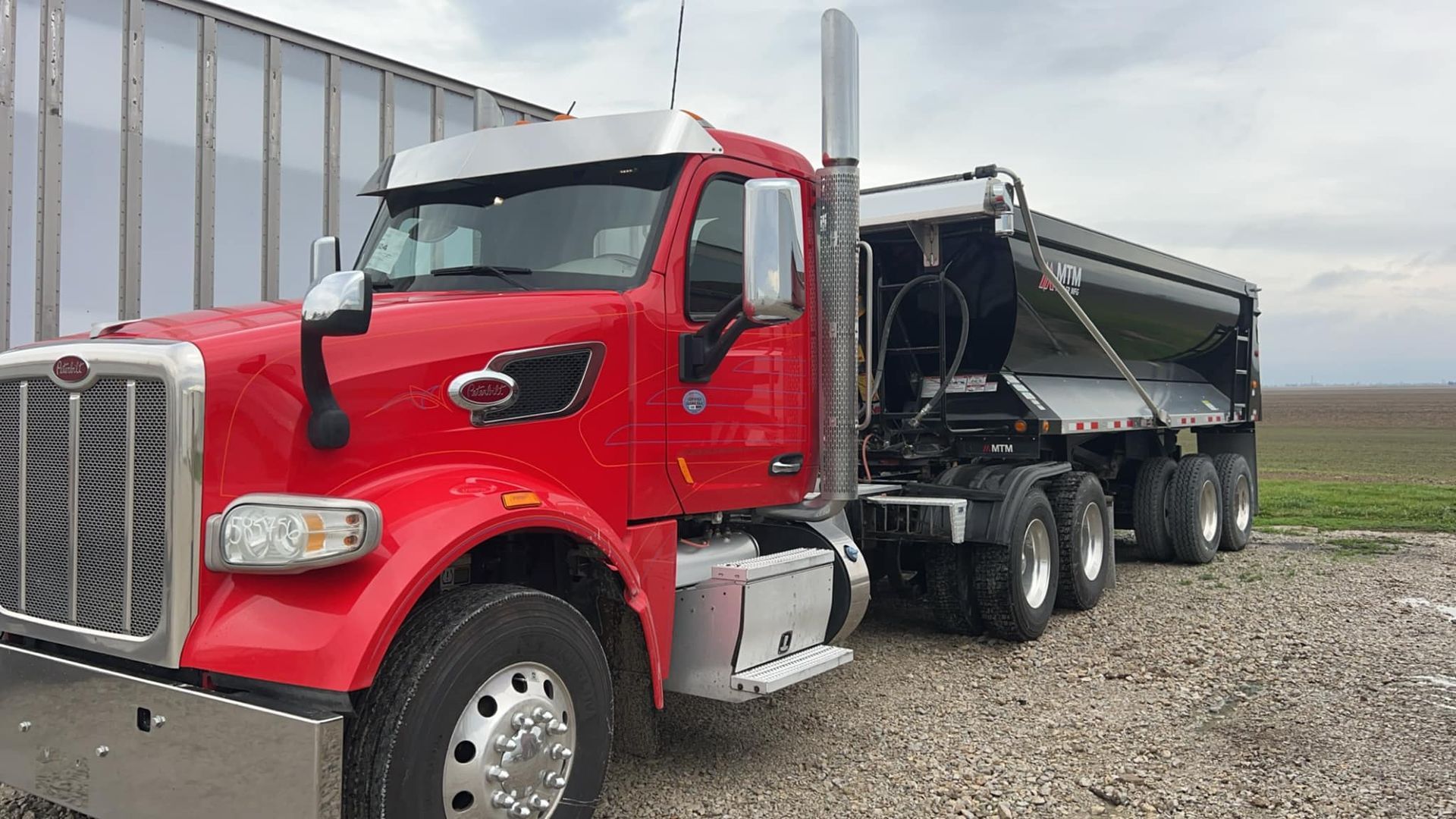 A red semi truck with a dump trailer attached to it is parked in a gravel lot.