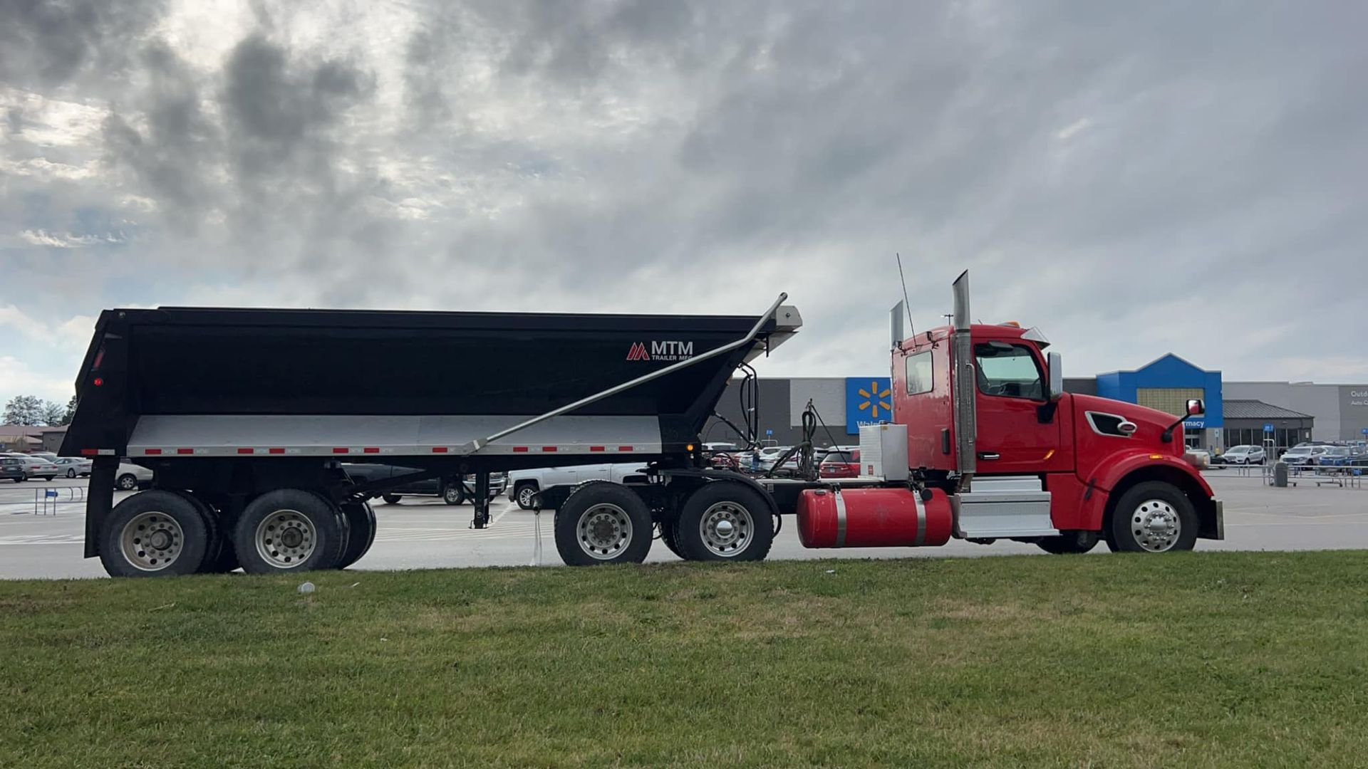 A red semi truck with a black dump trailer is parked in a grassy field.