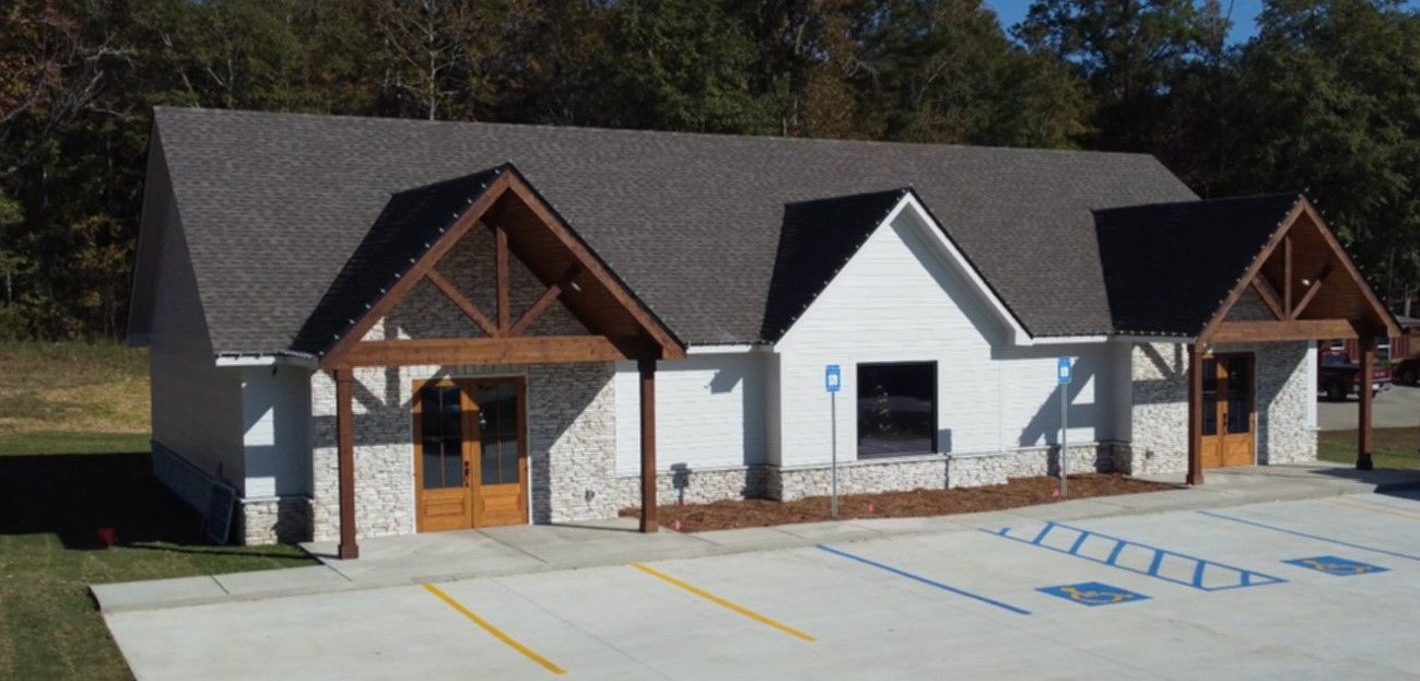 A white building with a gray roof and wooden accents. It has a parking lot and a wooded background.