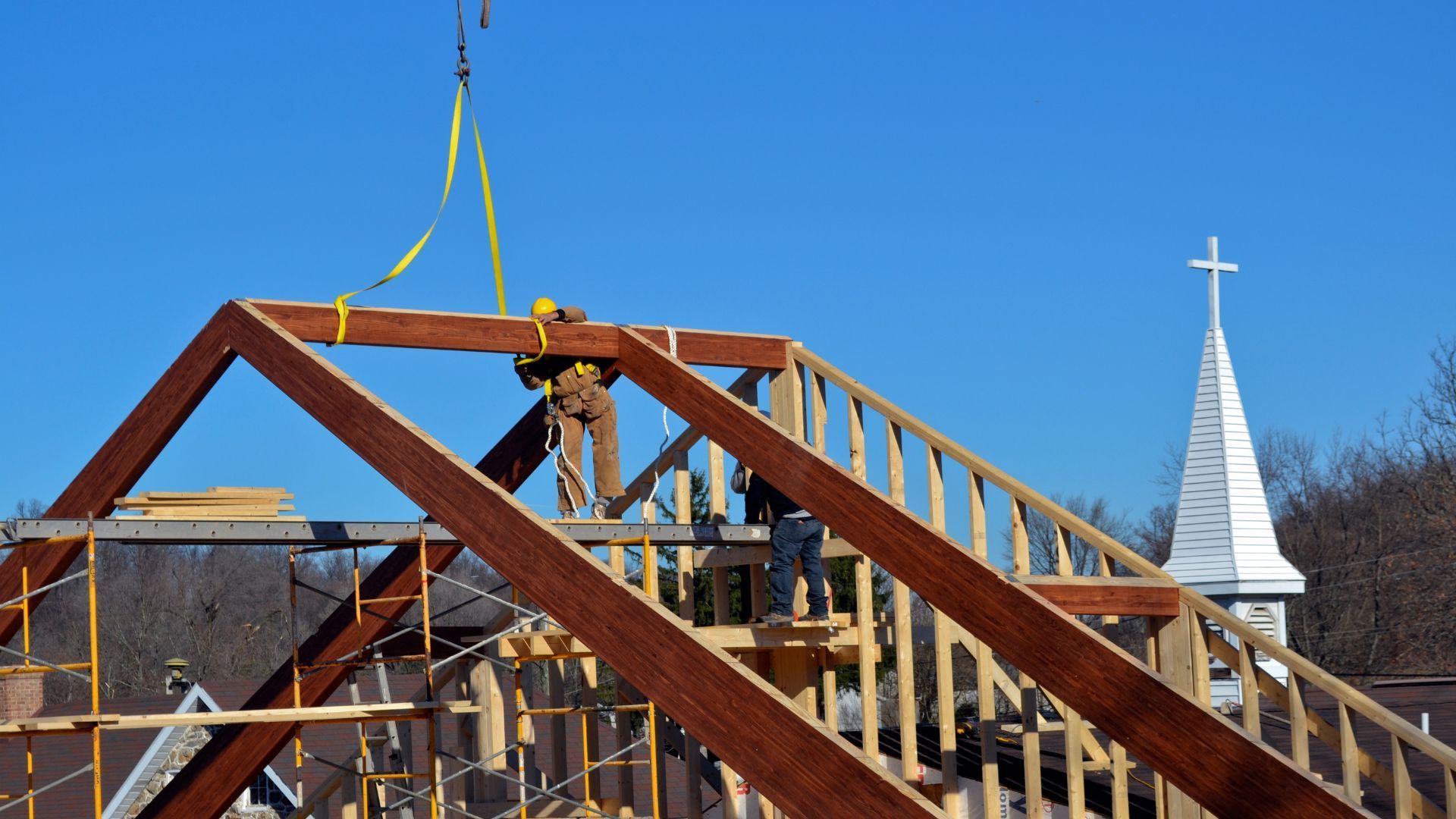 Construction workers install a roof truss on a building, crane in use, clear blue sky, white steeple in background.