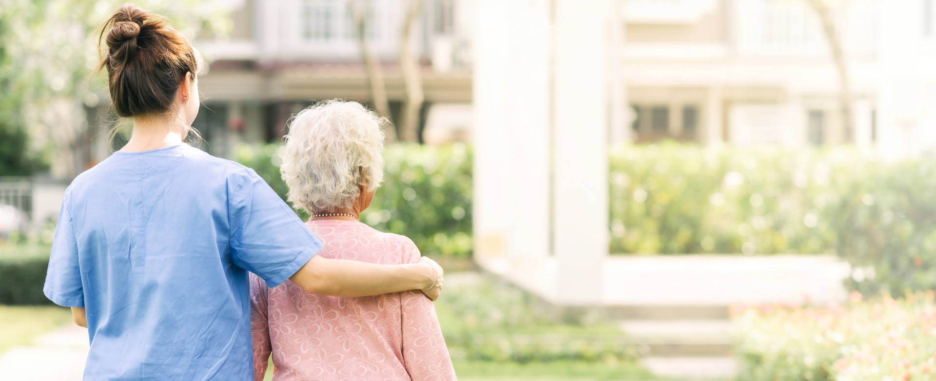 A caregiver and elderly woman walking with arms around each other outdoors.