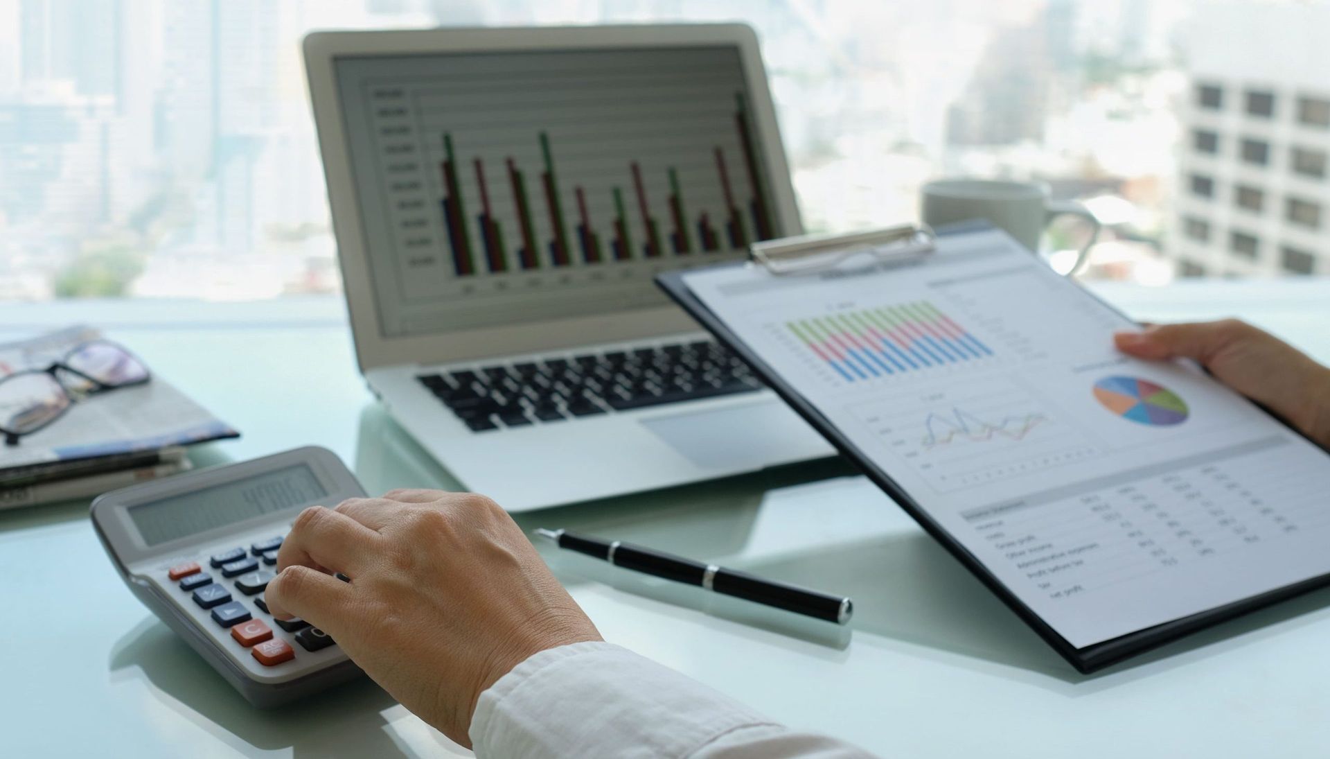 Person calculating numbers with a calculator, reviewing charts on a clipboard and laptop.  Office setting with cityscape view.