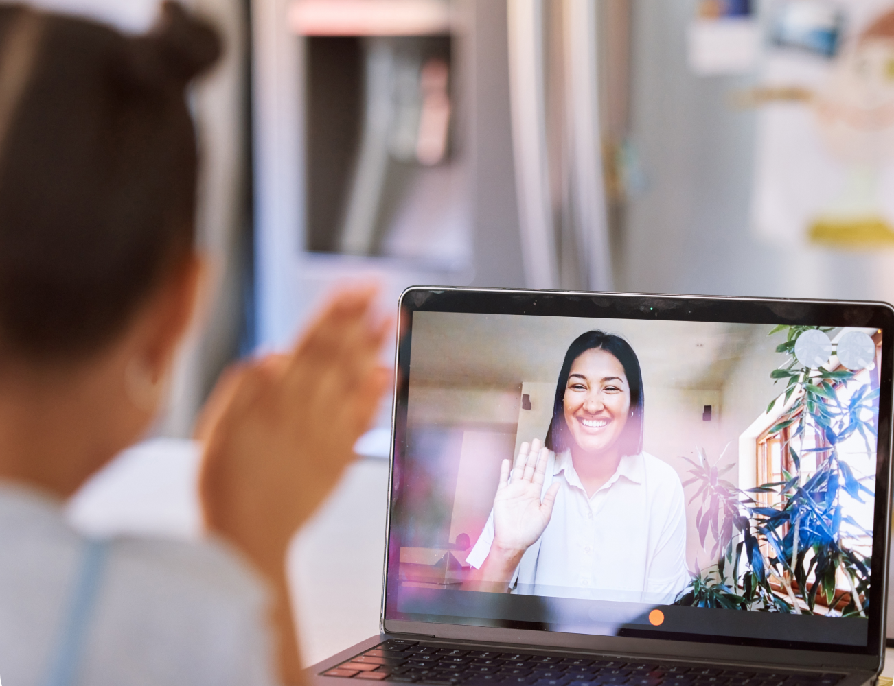 A young girl waves at a smiling woman on a laptop screen, inside a home with a fridge in the background.