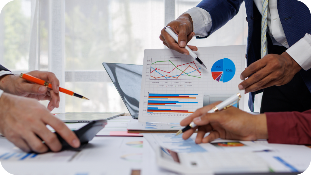 People in business attire reviewing charts and graphs during a meeting at a table, using pens to point at data.