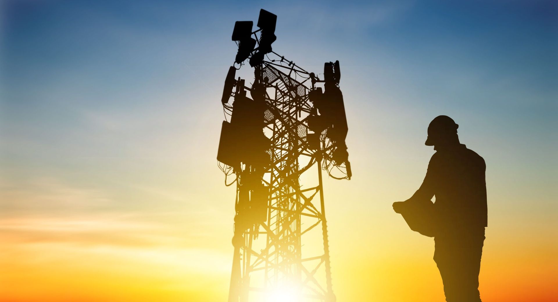 Silhouette of a worker in a hard hat inspecting a communications tower against a sunset.