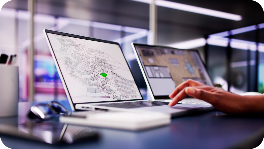 Two laptops on a desk, one displaying a map with a green marker. A person uses the other, blurring the hand in action.