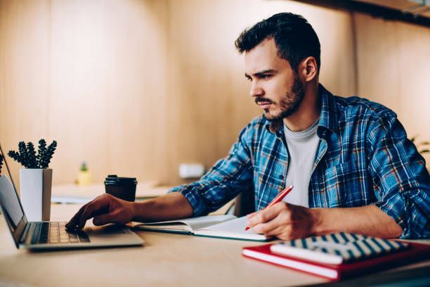 Man with beard in plaid shirt working on a laptop, taking notes in a notebook.