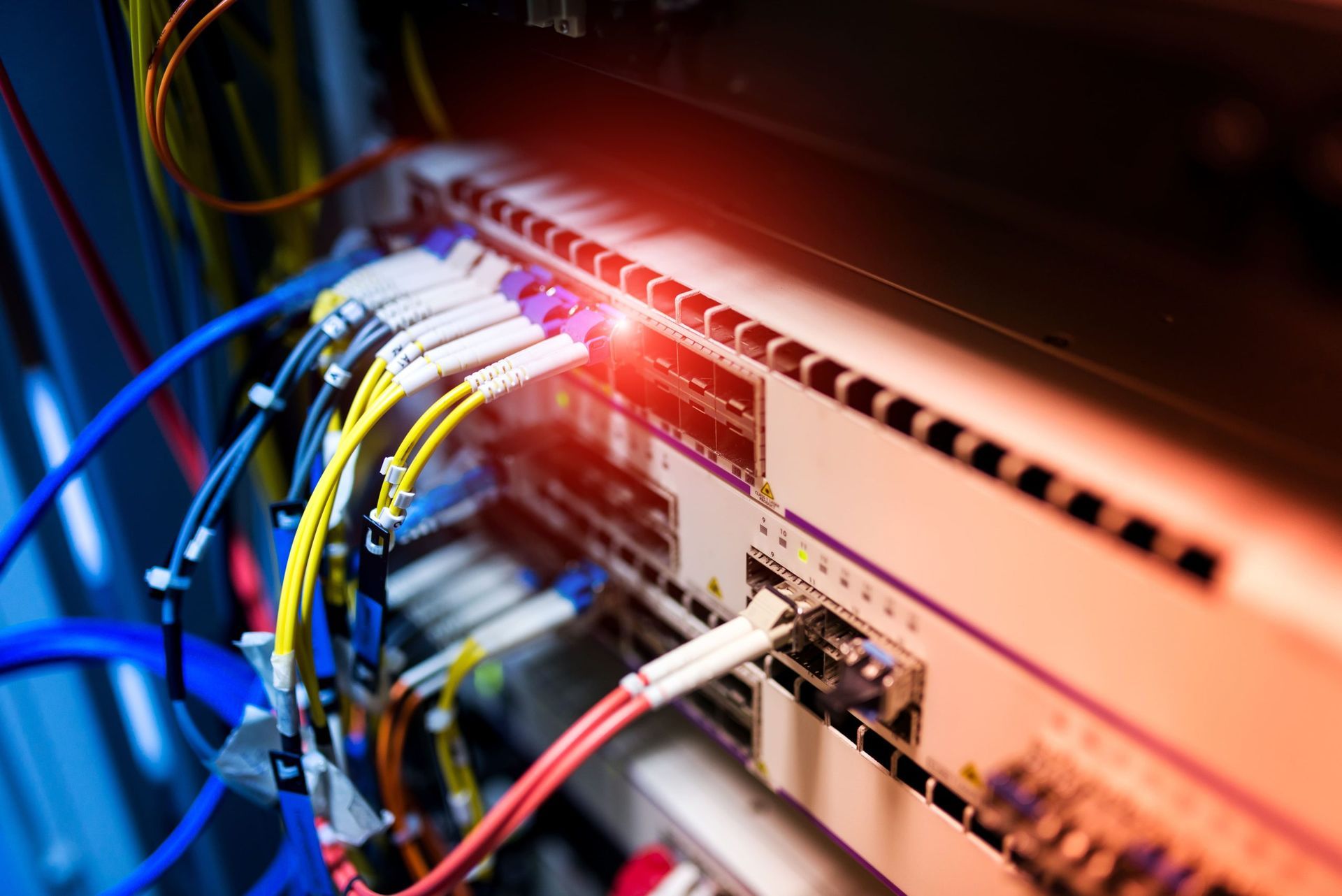 Close-up of a server rack with many colored cables connected to networking hardware, lit with red light.