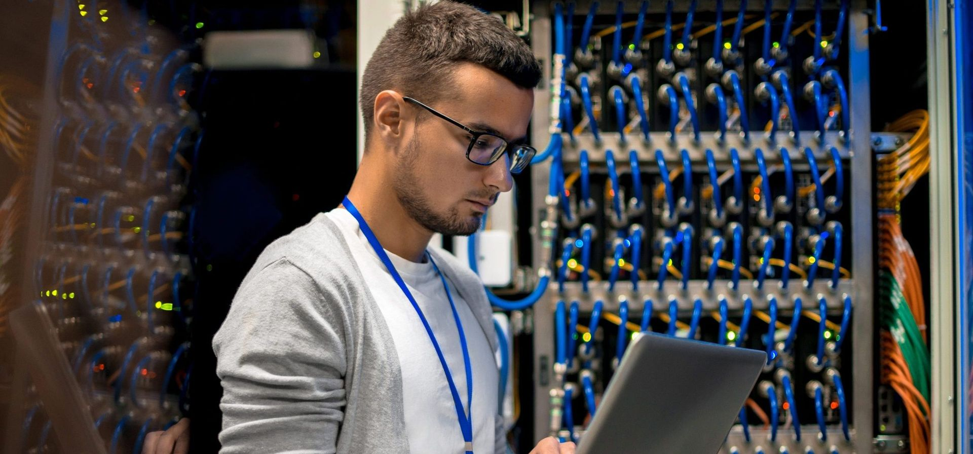 Man with glasses using a laptop in a server room, looking at servers, cable management.