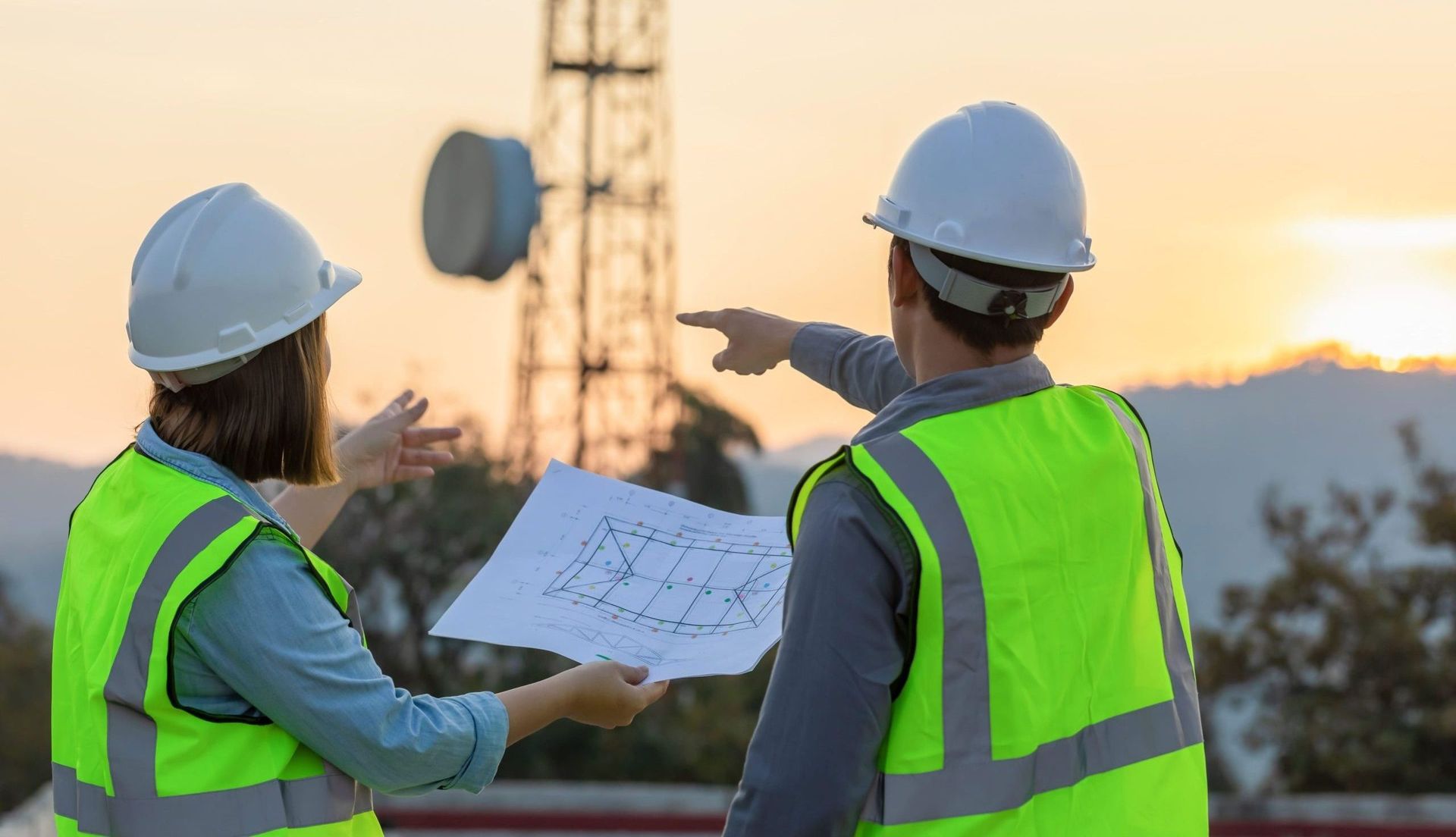 Two engineers in safety vests and hard hats examine a blueprint near a telecommunications tower at sunset. One points.