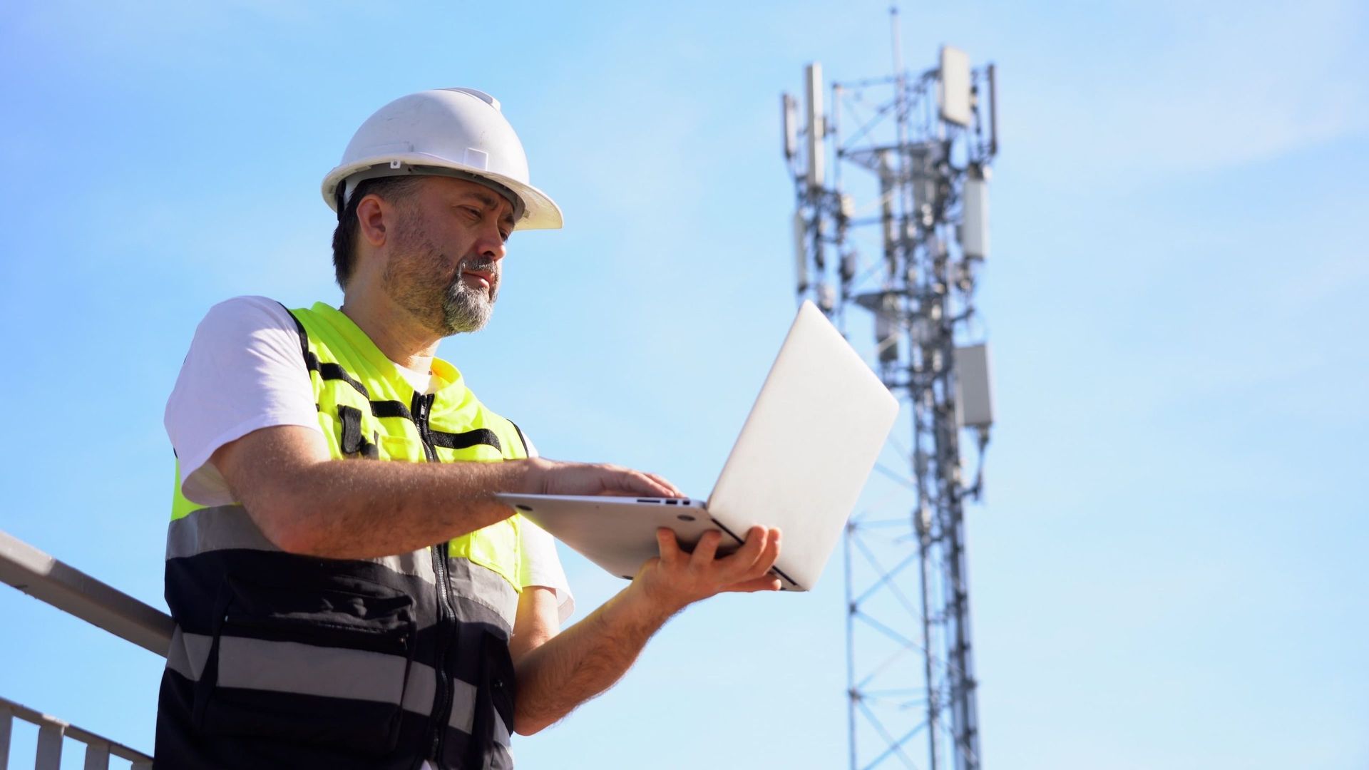 A telecom worker in a hard hat and vest using a laptop, with a cell tower in the background against a blue sky.